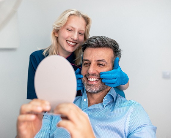 Man smiling at reflection in mirror with dentist touching his cheeks